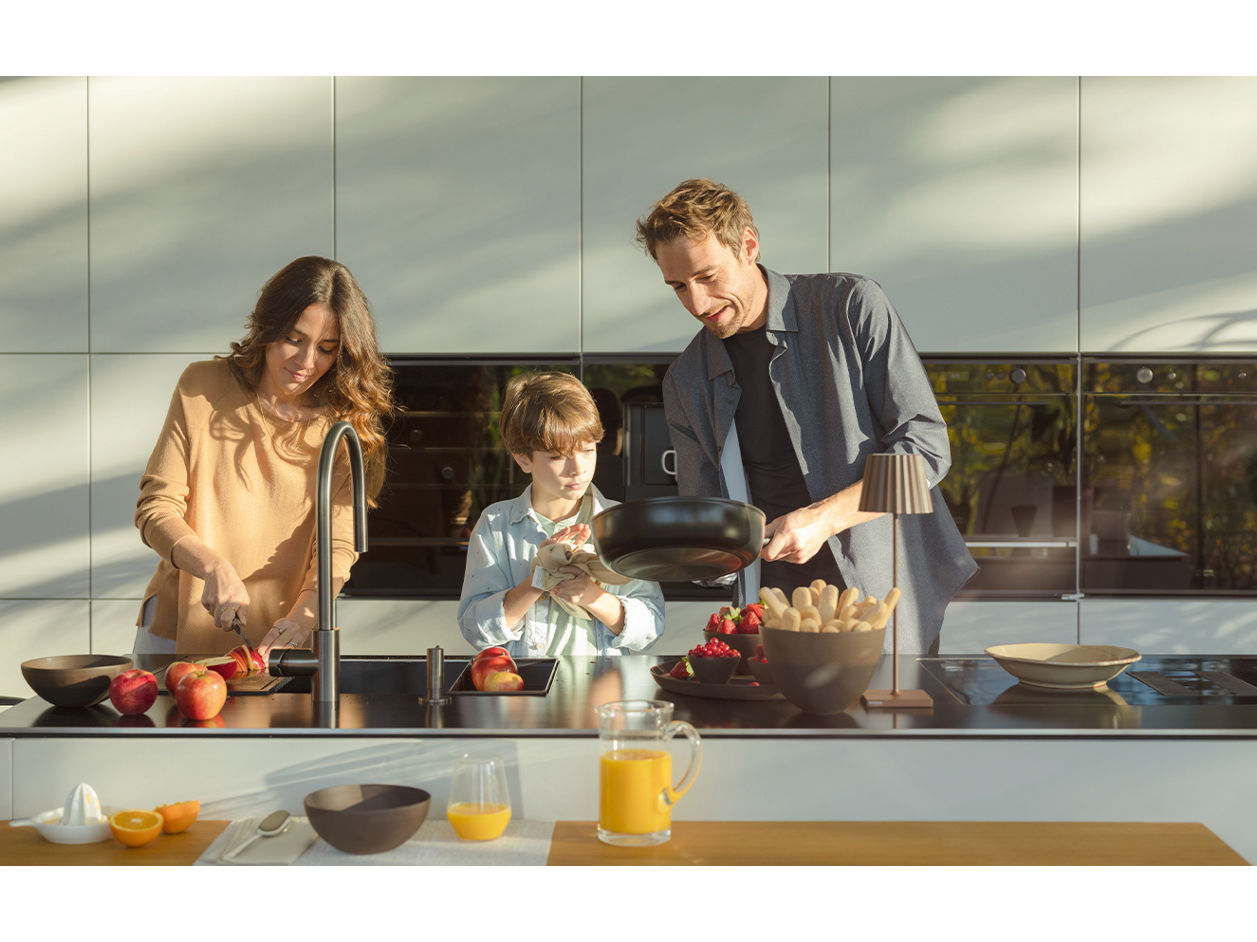 A family of three is in a modern kitchen. The woman is slicing fruit, the boy is holding a pot, and the man is pouring something into it. A pitcher of orange juice and a bowl of apples are on the counter. Sunlight streams in, casting shadows